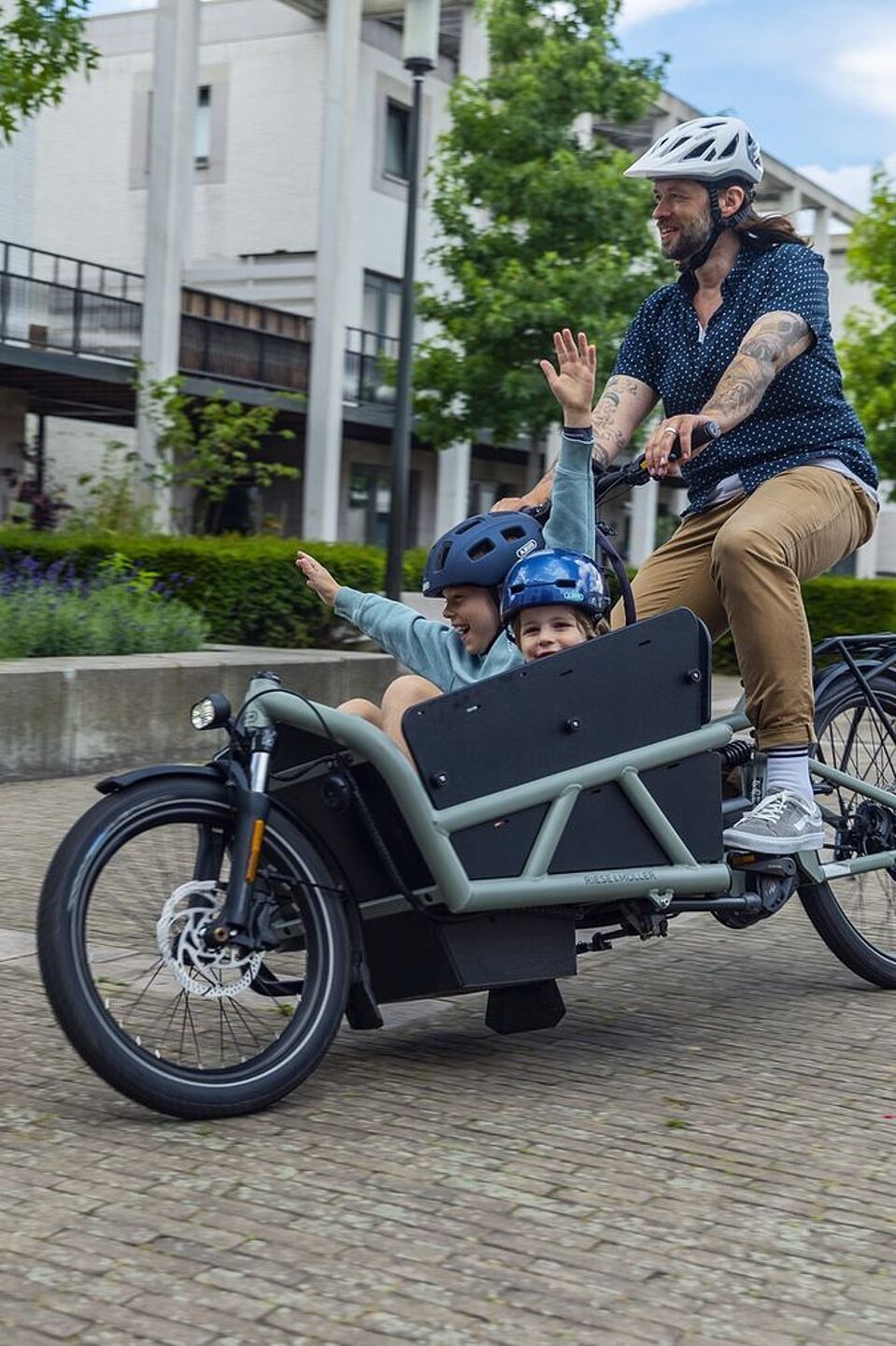 Kinder und Eltern haben Spaß, wenn sie gemeinsam Radfahren. Zwei Kinder mit Helm in einem Lastenrad jubeln, eine erwachsene Person mit Helm fahrt das Lastenrad