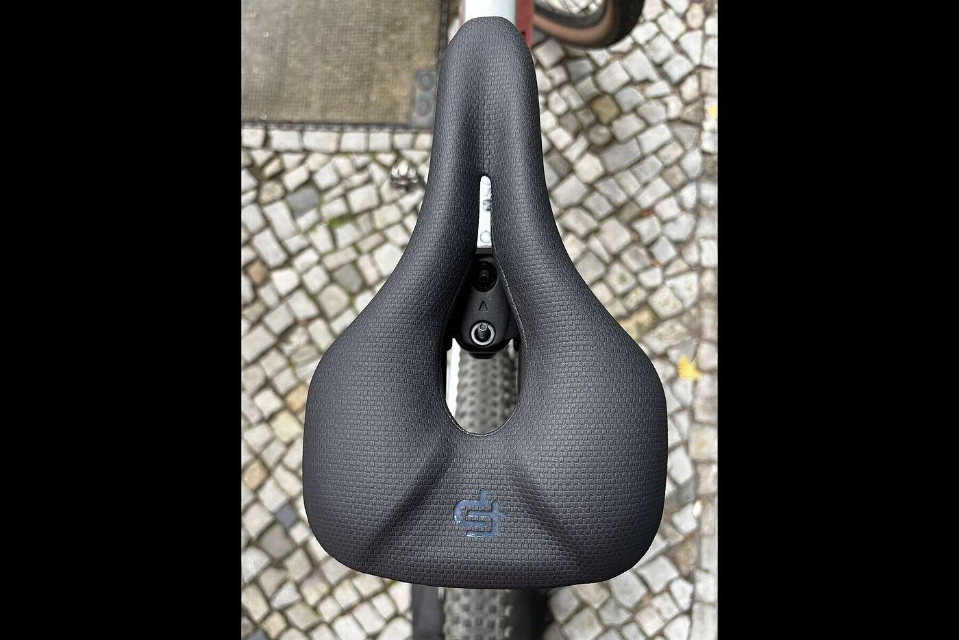 Fingerprint oben Schwarzer Fahrradsattel mit großer mittlerer Öffnung, von oben auf Kopfsteinpflaster fotografiert.