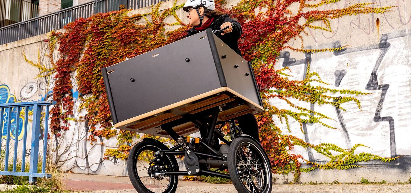 Chike Kurve Person mit Helm fährt ein dreirädriges Lastenrad mit großer Transportbox vor einer bewachsenen Wand.