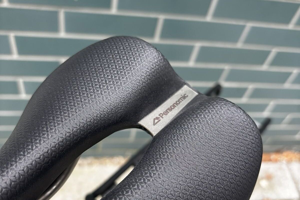 Personomic Detail Detail eines schwarzen Fahrradsattels mit feiner Struktur und Metallplakette vor hellgrüner Kachelwand.